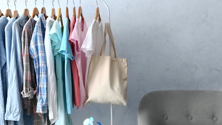 Mit unterschiedlicher heller Kleidung und beigem Jutesack behängte Kleiderstange vor blaugrauem Hintergrund, daneben Ausschnitt eines grauen Sofasessels, im Vordergrund blaue Orchidee