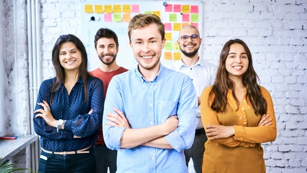 Porträt von fünf versetzt neben- und hintereinander stehenden Personen, die vor einer Ziegelmauer stehen. Hinter den Personen hängt an der Wand eine Tafel mit Zetteln