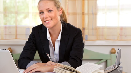 Portraitaufnahme einer Person, die an einem Schreibtisch sitzt, im Hintergrund Vorhänge. Sie tippt mit der linken Hand in einen Laptop, in der linken hält sie einen Kugelschreiber. Auf dem Holztisch befinden sich ein aufgeklapptes Buch und ein Telefon. 