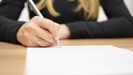 Eine Person unterschreibt mit einem Kugelschreiber auf einem bedruckten Blatt Papier