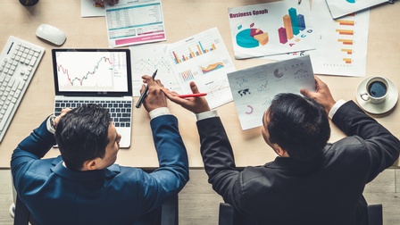 Zwei Personen in Businesskleidung werden aus der Vogelperspektive sitzend an einem Tisch gezeigt wie sie Statistiken am Papier und am Laptop besprechen, die Hände einer dritten Person ragen von der anderen Seite des Tisches herein