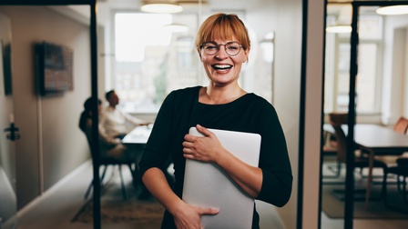 Lachende Person mit Brillen hält Notebook in Armen, im Hintergrund verschwommen Besprechungszimmer mit sitzenden Personen