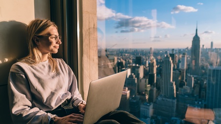 Sanft lächelnde Person mit Brillen sitzt an Fenster mit aufgeklappten Laptop am Schoß und blickt ins Sonnenlicht auf Stadtpanorama mit Hochhäusern