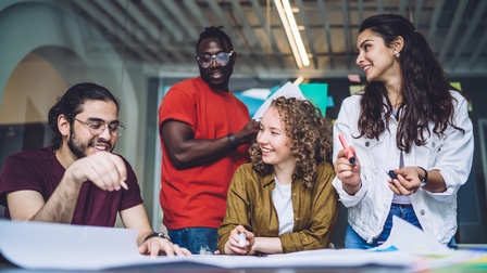 Zwei Personen sitzen, zwei Personen stehen an einem Tisch. Vor ihnen ist ein großes Plakat. Eine Person blickt darauf. Eine Person hält einen Zettel. Zwei weitere Personen halten einen Stift
