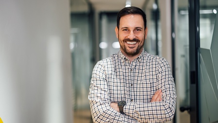 Lächelnde Person mit dunklen kurzen Haaren, Bart und kariertem Hemd steht mit verschränkten Armen in einem Flur und blickt Richtung Kamera