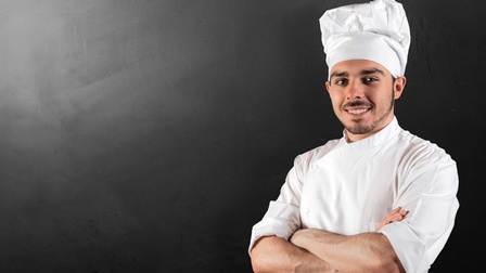 Porträt einer Person mit Kochschürze und Kochmütze vor dunkelgrauem Hintergrund mit verschränkten Armen