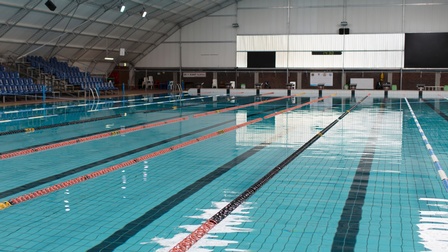 Ein leeres Hallenbad mit einem breiten und langen Pool, das durch rot-schwarze, schwimmende Linien in mehrere Bahnen unterteilt ist. Am linken Rad stehen mehrere blaue Sesselreihen