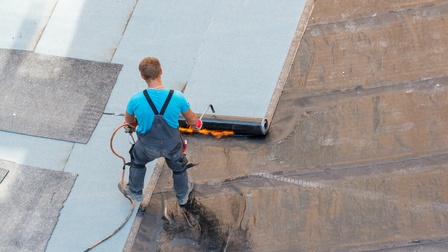 Aufnahme einer Person aus der Vogelperspektive, die auf einem Flachdach steht und mit einem Gerät das Dach versiegelt. Die Person trägt Handschuhe