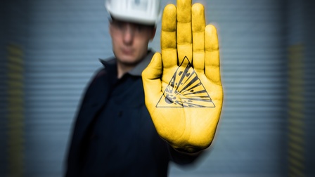 Eine Person mit Helm hält ihre gelb angemalte Hand in die Kamera. Auf der Handinnenfläche ist ein Dreieck, in dem eine Explosion ist