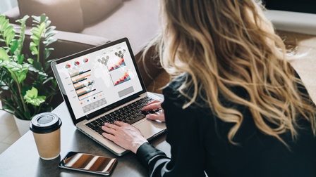 <br />
Person mit Bluse sitzt vor einem Laptop an einem Schreibtisch in einem Wohnzimmer mit Grünpflanze und blickt auf Grafiken, Diagrammen und Statistiken während daneben ein Coffee-to-go Becher sowie ein Smartphone liegt