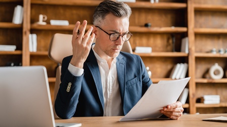Person im Anzug trägt eine Brille und sitzt mit fragendem Blick an einem Schreibtisch mit Computer und hält Papier in einer Hand ein Papier und in der anderen einen Stift, im Hintergrund steht ein braunes Holzregal mit Büchern und Dekoelementen