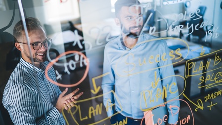Zwei Personen stehen hinter transparentem Board mit bunten Schriftzügen