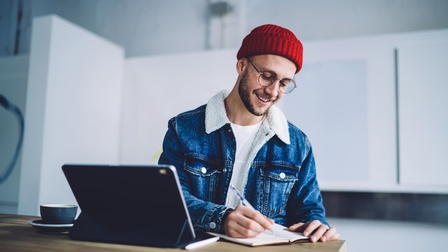 Person mit roter Mütze, Bart und Brille sowie Jeansjacke steht bei einem Holztisch und macht Notizen in einem Heft, daneben steht ein Tablet sowie eine Tasse, im Hintergrund zeigt sich eine weiße Einrichtung
