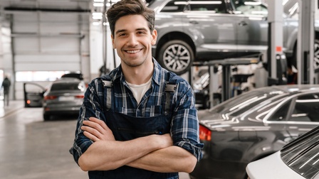Porträt einer lächelnden Person in einem Overall und mit verschränkten Armen, die in einer großen Halle steht, in der mehrere Fahrzeuge stehen