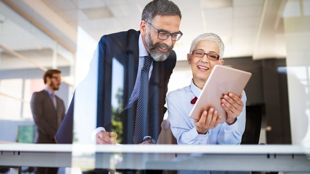 Zwei Personen mit Brille und kurzen Haaren sowie Businesskleidung blicken freudig auf ein Tablet, im Hintergrund zeigt sich eine Bürosituation und eine weitere Person steht im Hintergrund