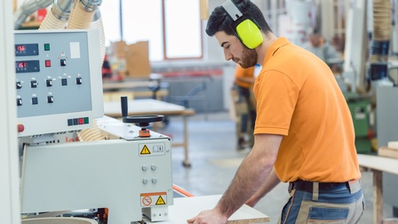 Person mit Hörschutz steht an Furniermaschine und hält Brett in Händen