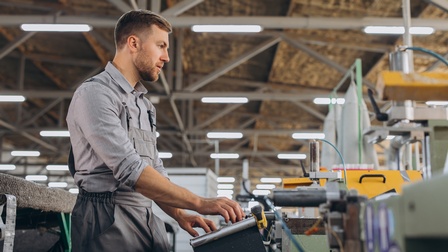 Seitliche Aufnahme einer stehenden Person, die ihre Hände auf einer metallenen Vorrichtung hat, mit der er eine Maschine bewegen kann, in einer großen Halle mit Deckenlampen.