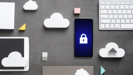 Mehrere Gegenstände nebeneinander auf grauem Hintergrund drapiert: Weiße Computertastatur, Smartphone auf dessen Display weißes Sicherheitsschloss leuchtet, weiße Modellwolken aus Schaumstoff, Tablet und weitere Formen aus Schaumstoff