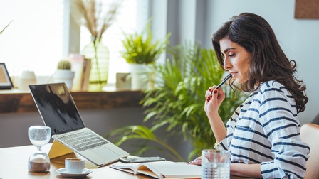Seitliche Aufnahme einer an einem Schreibtisch sitzenden Person. Vor ihr ist ein aufgeklapptes Buch, auf das sie blickt. Auf dem Tisch sind ein Laptop, eine Tasse, ein Tablet, eine Sanduhr und ein Glas. Im Hintergrund sind Topfpflanzen