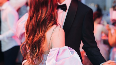 Zwei Personen tanzen eng miteinander. Die linke Person trägt ein helles Oberteil mit Puffärmeln sowie hat lange, offene, rötliche Haare. Die andere Person trägt einen Smoking mit Fliege