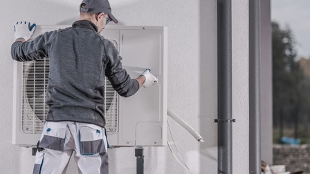Person in Rückenansicht mit Kappe und Arbeitshandschuhen befestigtWärmepumpen-Außengerät an weißer Hausfassade