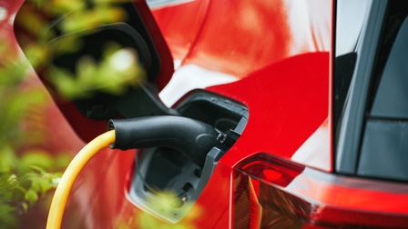 Nahaufnahme eines geöffneten Tankdeckels eines roten Elektro-Autos, aus dem ein dickes Stromkabel kommt. Im linken Bildrand sind verschwommene, grüne Blätter.