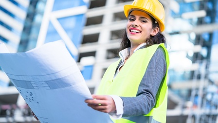 Porträtaufnahme einer Person, die einen Plan in der Hand hält, auf dem die Skizze eines Gebäudes zu sehen ist. Im Hintergrund sieht man verschwommen Hochhäuser. 