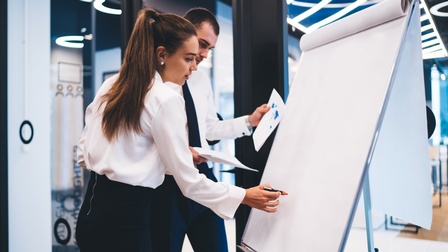 Zwei Personen in Bürokleidung stehen nebeneinander an einem Whiteboard. Die rechte Person notiert etwas mit einem Stift darauf. Die linke Person hält verschiedene Zettel