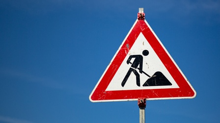 Ein dreieckiges Schild, auf dem die Silhouette einer Person eine Schaufel hält und sich nach vorne beugt. Vor ihr liegt ein Haufen. Dahinter ist der blaue Himmel