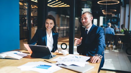 Zwei Personen in dunklen Anzügen sitzen in Besprechungszimmer vor Tablet, am Tisch liegen Unterlagen