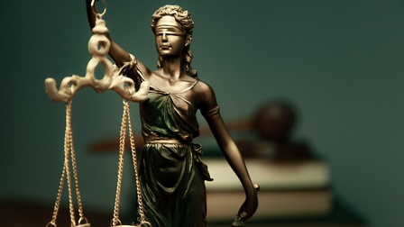 Statue Justitia, im Hintergrund sind Bücher sowie ein Richterhammer in der Unschärfe platziert