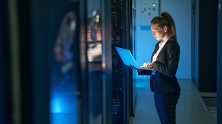 Person in Businesskleidung steht in einem Serverraum und hält einen Laptop in der Hand