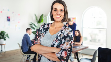 Lächelnde Person mit schulterlangen braunen Haaren steht mit verschränkten Armen und einer floralen Bluse in einem Büro während im Hintergrund mehrere Personen bei einem Schreibtisch sitzen