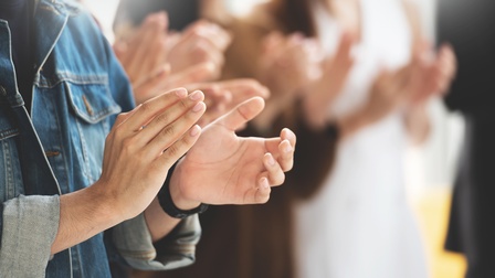 Nahaufnahme der Hände einer Person, die gerade klatscht. Im Hintergrund sind weitere Personen in Unschärfe fotografiert, die klatschen