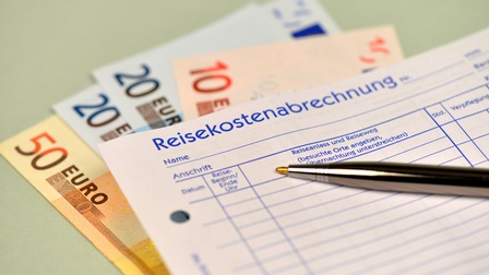 Detailansicht eines Formulars mit blauer Beschriftung Reisekostenabrechnung, darauf liegend Kugelschreiber, darunter liegend Geldfächer mit 50-, 20- und 10-Euro-Scheinen