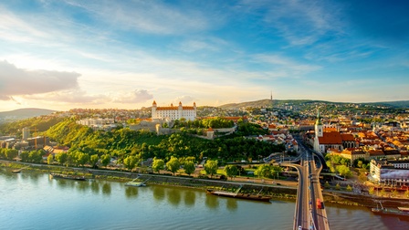 Vogelperspektive einer mehrspurigen Brücke, die über einen Fluss führt. Am Ende der Brücke ist am Ufer eine Stadt mit mehreren Gebäuden. Auf einem Hügel ist eine helle Burg. Am Ufer sind verschiedene Boote