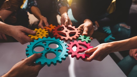 Sechs Hände von fünf Personen halten sechs unterschiedlich große Zahnräder in die Mitte. Teilweise berühren sich die Zahnräder