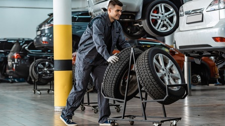 Lächelnde Person schiebt zwei Autoreifen auf Rollgestell durch Autowerkstatt