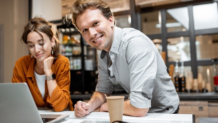 Zwei Personen stehen in einem Lokal hinter einem Tresen. Die linke Person blickt auf einen aufgeklappten Laptop, der auf dem Tresen steht. Die rechte Person blickt direkt in die Kamera. Am Tresen steht ein Kaffeebecher
