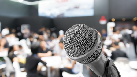 Nahaufnahme eines Mikrofons vor einer Halle, in der Personen in Gruppen an Tischen sitzen