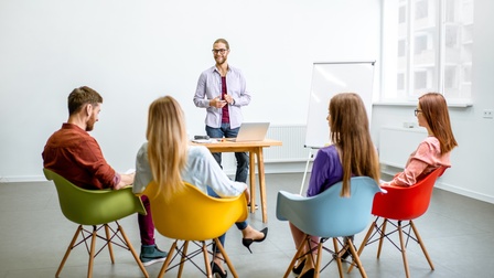 Rückenansicht von vier Personen, die in Sesseln nebeneinander sitzen. Den Personen gegenüber steht eine weitere Person hinter einem Tisch. Neben der Person ist ein Whiteboard.