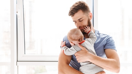 Frontalaufnahme einer Person mit kurzen Haaren und Bart, die vor einem Fenster steht. Mit beiden Händen hält die Person ein Baby vor den Oberkörper und blickt darauf