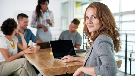 Person mit längeren rötlichen Haaren blickt in die Kamera, während sie bei einem aufgeklappten Laptop sitzt, im Hintergrund stehen und sitzen weitere Personen bei einem Besprechungstisch