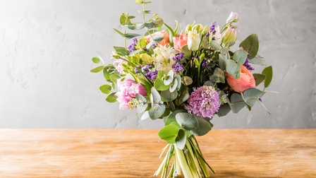Zusammengebundene, bunte Blumen mit langen grünen Stielen stehen auf einer Holzplatte. Die Mauer im Hintergrund ist grau