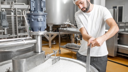 Person mit Haarnetz rührt großen Topf mit gestockter Milch