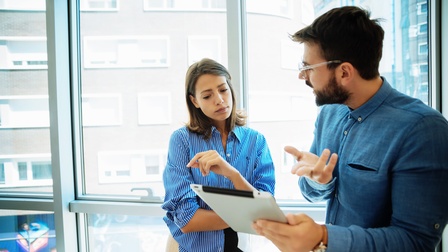 Eine Person mit kurzen Haaren, Bart und Brille hält ein Tablet in Richtung einer weiteren Person mit der sie zugewandt spricht, die zweite Person blickt interessiert auf das Tablet, im Hintergrund zeigt sich eine vollflächige Glasfront