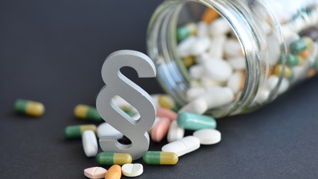 Fokus auf weißes Paragrafenzeichen platziert zwischen verschiedenfarbigen und -förmigen Pillen, die aus vollem umgekippten Rexglas kullern auf grauem Untergrund