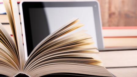 Im Vordergrund ist ein aufgeklapptes Buch. Dahinter lehnt ein Tablet an einem Bücherstapel