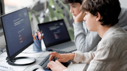 Seitliche Aufnahme zweier Teenager an einem Tisch. Auf dem Tisch steht ein Bildschirm, auf den beide blicken. Daneben sind Kopfhörer, verschiedene Stifte und ein Laptop