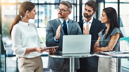 Vier Personen in Bürokleidung stehen nebeneinander an einem Stehtisch, auf dem ein Laptop steht. Die Person am Laptop gestikuliert mit den Händen, die anderen blicken zu ihr. Im Hintergrund sind Fensterfronten